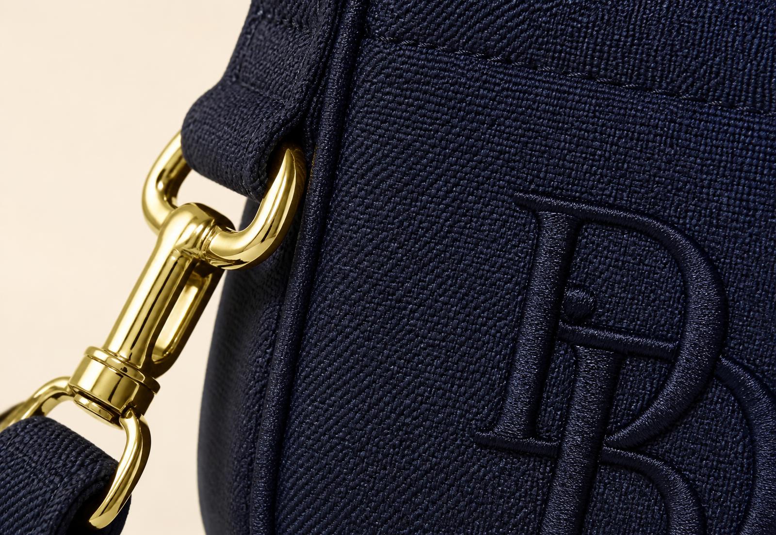 Detailaufnahme einer marineblauen CAMELLIA-Stofftasche mit goldenem Karabiner, gewebtem Gurtband und gestickter Monogramm-Stickerei
